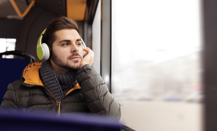 Junger Mann sitzt mit Winterjacke und Kopfhörern in einem Bus und schaut verträumt aus dem Fenster.