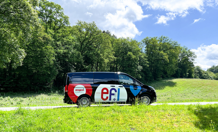 Ein Mini-Van mit dem Aufdruck "efi" fährt durch eine grüne, ländliche Landschaft.