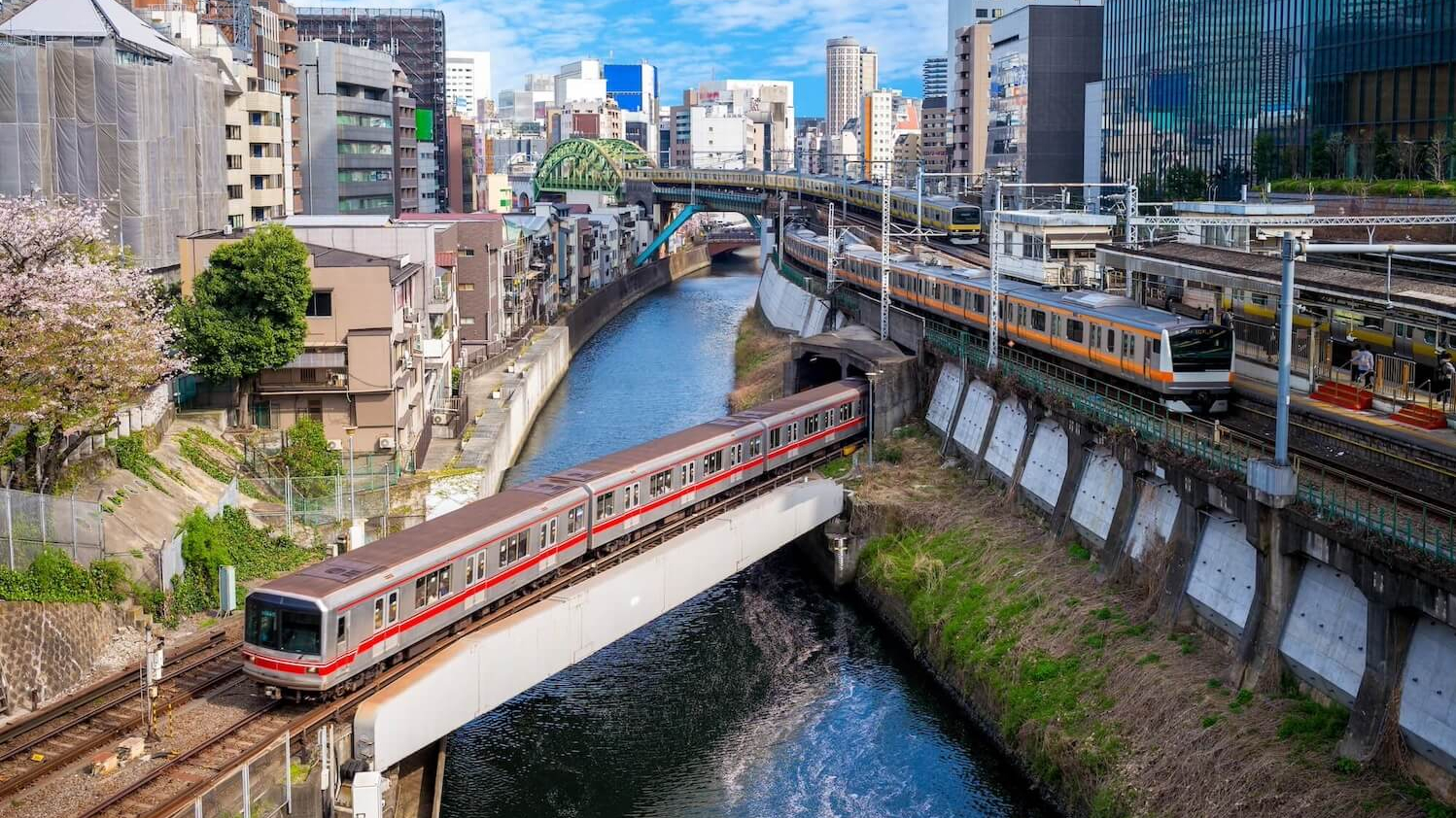 Eine rote Bahn fährt von rechts hinten nach links vorne über eine Brücke über einen Fluss. Drumherum ist eine belebte Stadt.