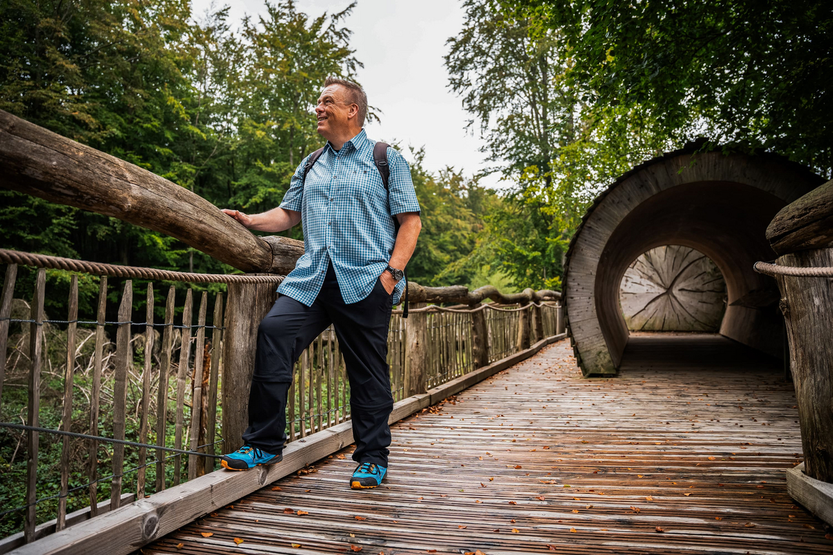 Ein Mann in blauem Outfit steht auf einem Wanderweg an einer hölzernen Brüstung. Um ihn herum ist Wald.