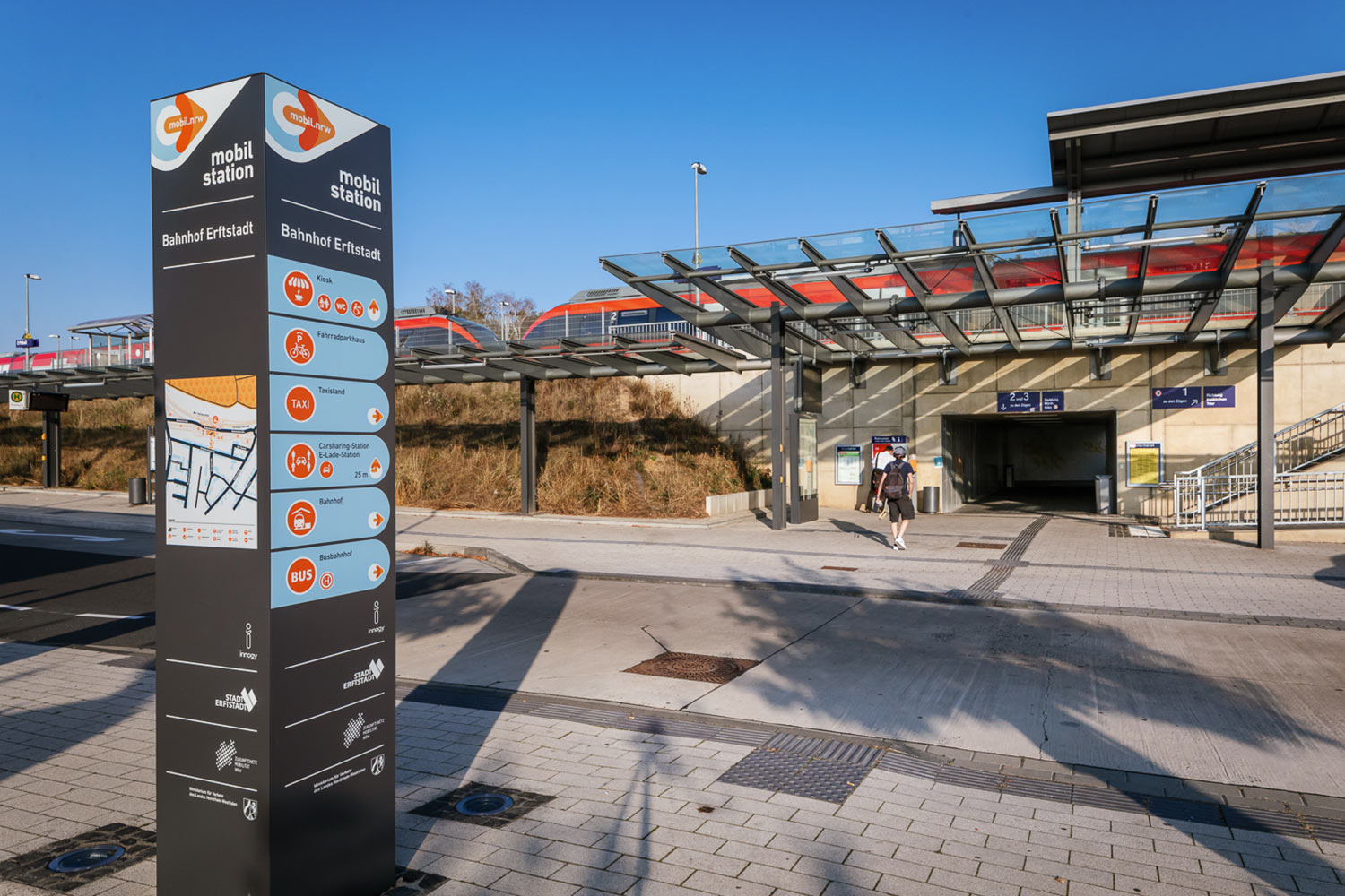 Die Säule der Mobilstation am Bahnhof Erftstadt. Im Hintergrund fährt ein Zug vorbei.