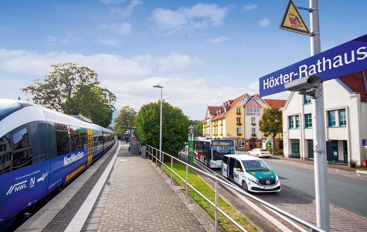 Ein Zug steht an einem Bahngleis, auf der Straße daneben parken ein Holibri-Fahrzeug und ein Linienbus.