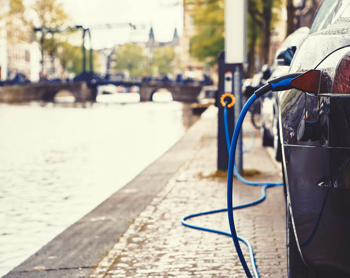 Teilausschnitt eines E-Autos, an dem ein Ladekabel hängt, rechts im Bild. Im linken Teil des Bildes sieht man auf die Grachten in Amsterdam.