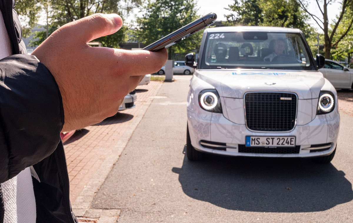 Ein Mann hält ein Handy in der Hand. Ein LOOPmünster-Fahrzeug komt auf ihn zu.