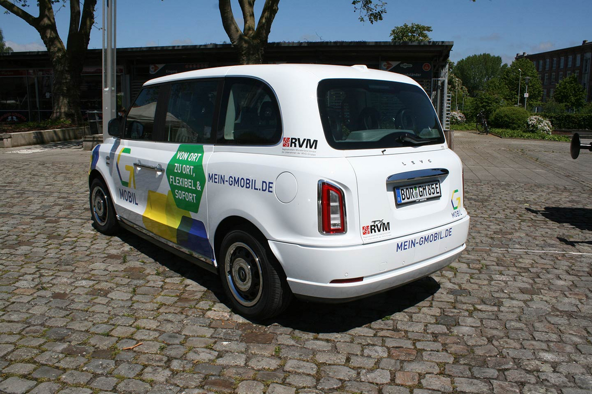 Ein kleines weißes Auto steht auf einem Kopfsteinplatz. Auf das Auto wurden gelbe und blaue Sechsecke lackiert, außerdem sind das Logo von G-Mobil und RVM abgebildet.