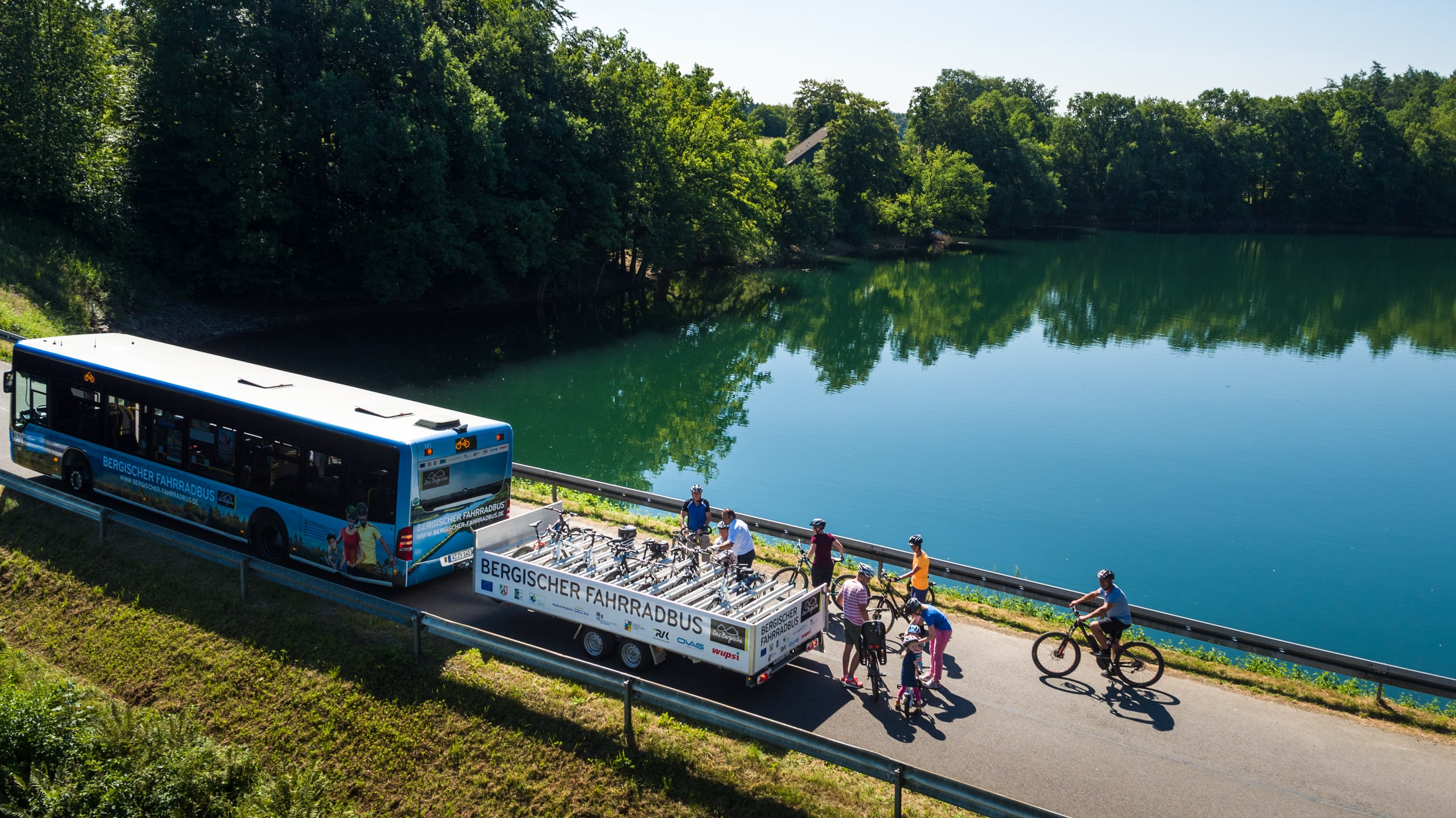 An einer Talsperre stehen Menschen neben einem Bus mit Fahrradanhänger.