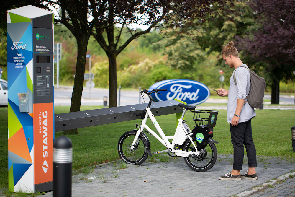 Ein junger Mann steht vor einem E-Bike an einer Verleihstation und schaut auf sein Handy.