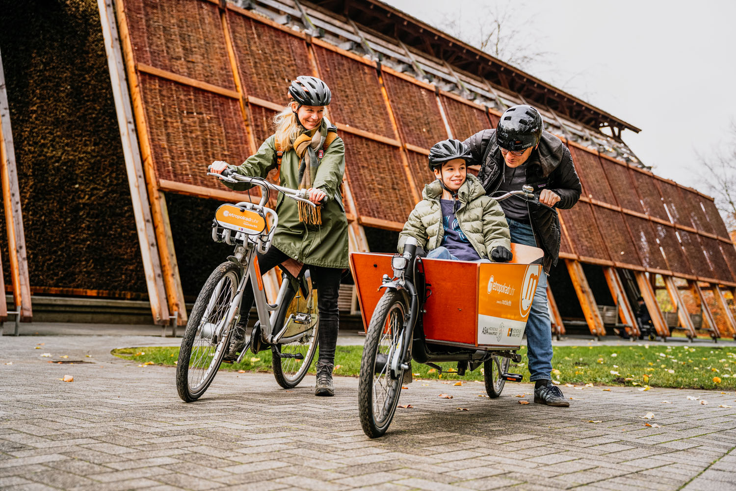 Eine Familie mit Vater, Mutter, Sohn fährt mit einem Leihrad und einem geliehenen Lastenrad durch die Natur. Der Sohn sitzt vorne in der Box des Lastenrads.