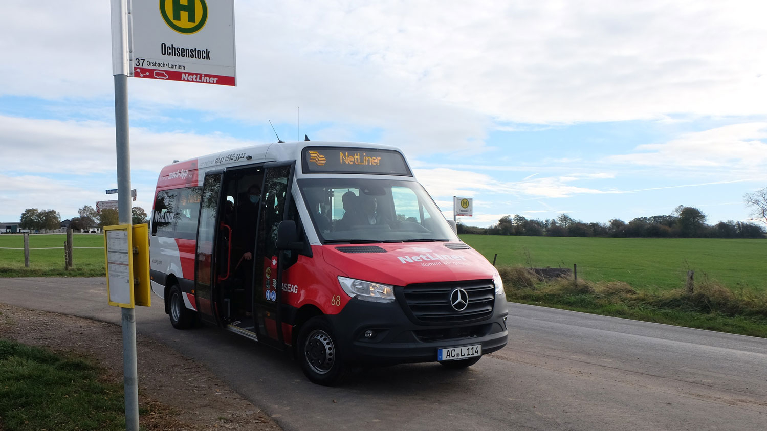 Ein rot-weißer Kleinbus steht vor einem Feld an einer Haltestelle.