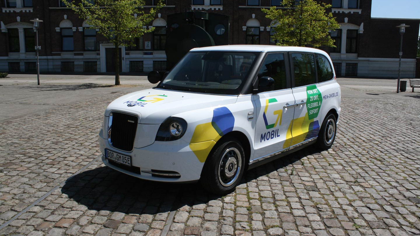 Ein kleines weißes Auto steht auf einem Kopfsteinplatz. Auf das Auto wurden gelbe und blaue Sechsecke lackiert, außerdem ist das Logo von G-Mobil zu sehen.