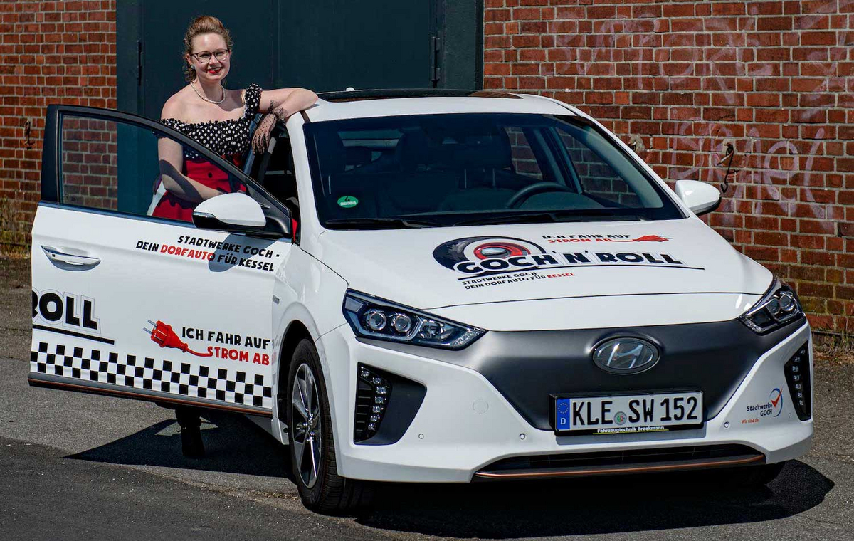 Eine Frau im Rockabilly-Style steht neben einem weißen E-Auto vor einer Backsteinmauer.