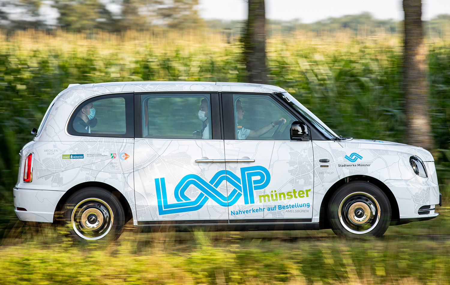 Ein LOOPmünster Fahrzeug fährt eine Straße entlang, dahinter sind grüne Büsche und Bäume zu sehen.