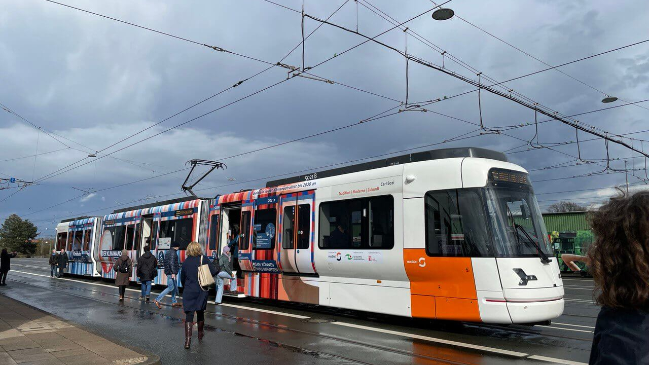 Eine weiß-orangene Straßenbahn mit Menschen vor grau-blauem Himmel.