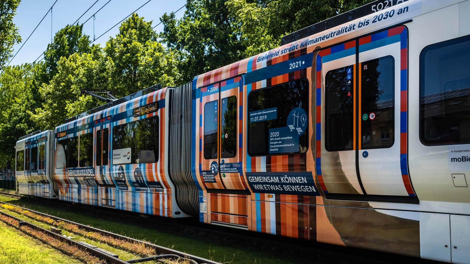 Eine rot-blaue gestreifte Straßenbahn fährt durchs Grüne.