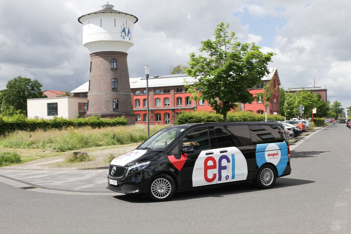 Ein schwarzer Minivan mit dem rot-blau-weißen Aufdruck "efi" steht auf einer Straße. Im Hintergrund: Ein Turm und Häuser.