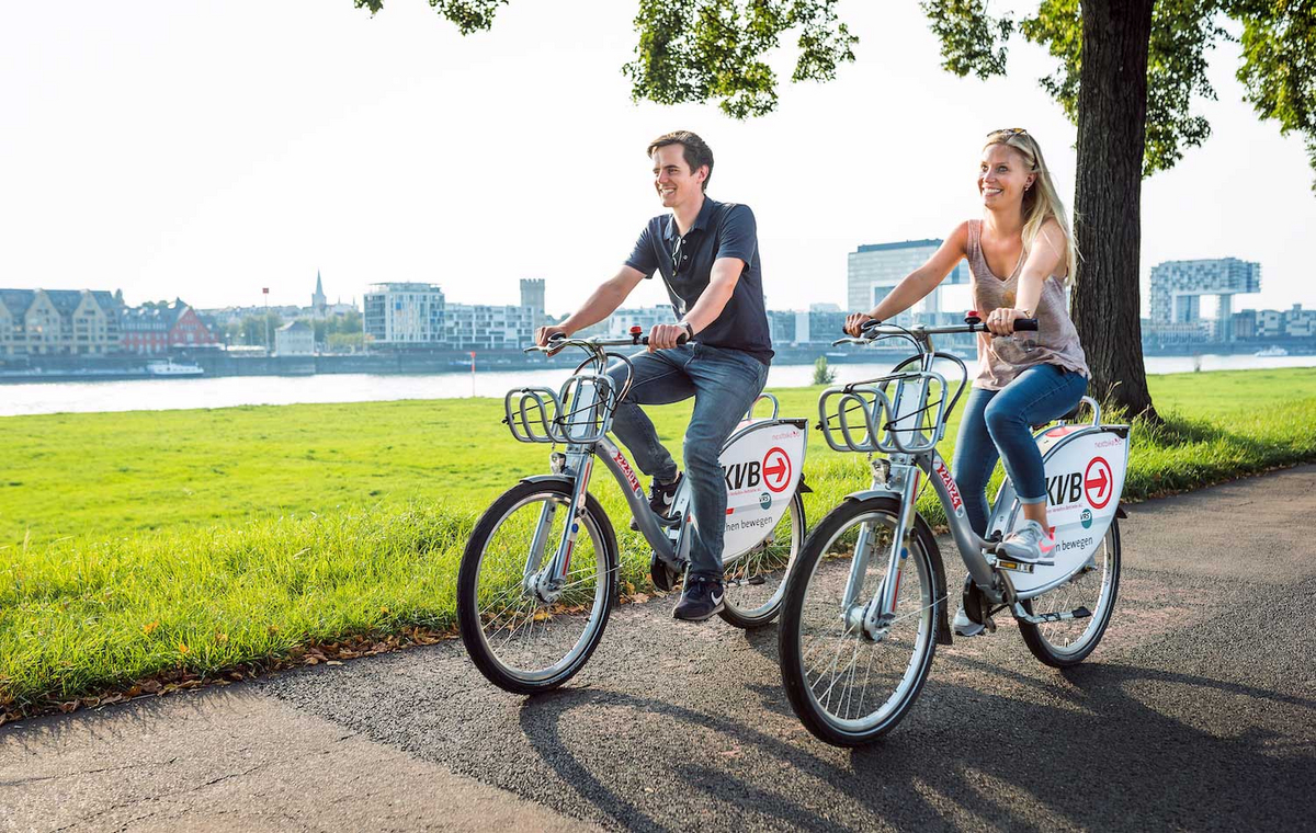 Ein Mann und eine Frau fahren auf Leihrädern über einen natürlichen Weg am Rhein in Köln entlang.