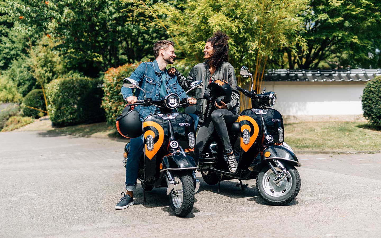 Ein junger Mann und eine junge Frau sitzen auf zwei orange-schwarzen E-Rollern mit der Aufschrift Alma.