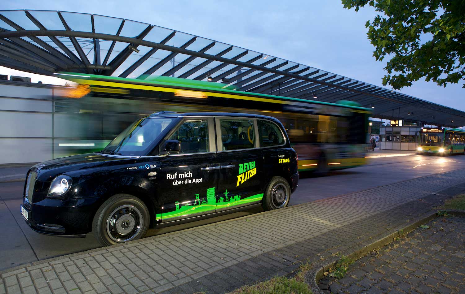 Ein kleines schwarzes Auto mit dem Aufkleber Revierflitzer steht an einer Haltestelle, im Hintergrund fahren gelb-grüne Busse.